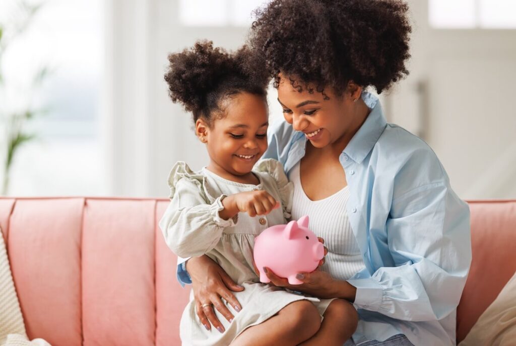 Mother and daughter putting a coin into a pink piggy bank at home.